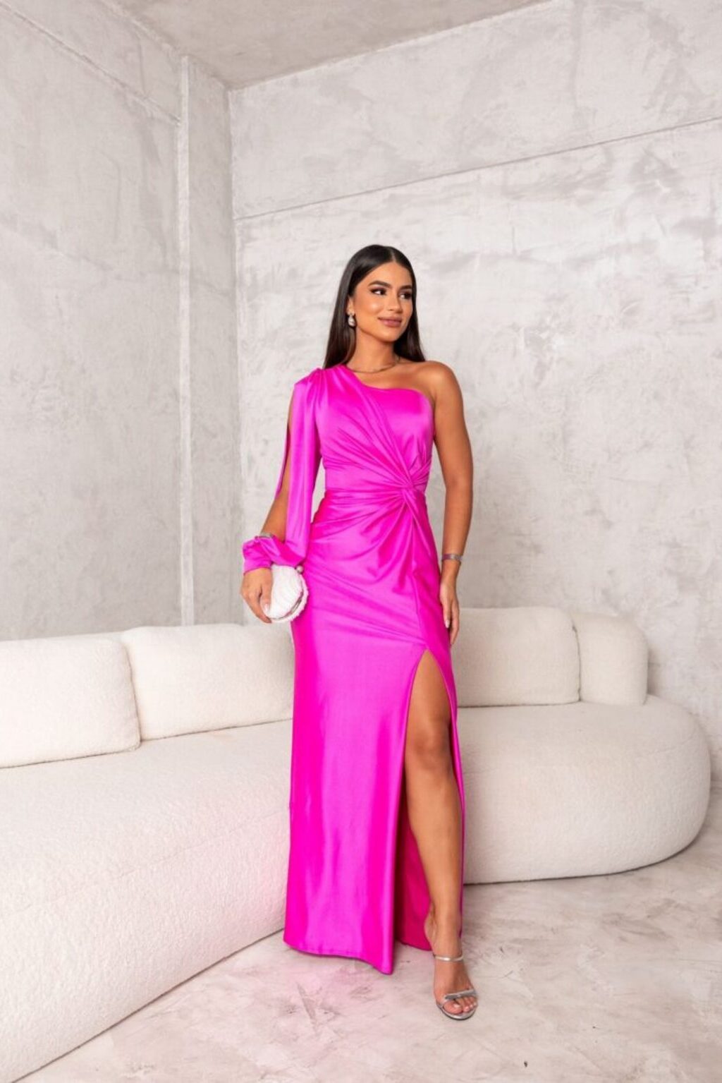 Donna in abito rosa acceso monospalla con spacco, su divano bianco in stanza moderna. Eleganza, moda femminile, abbigliamento da sera.
