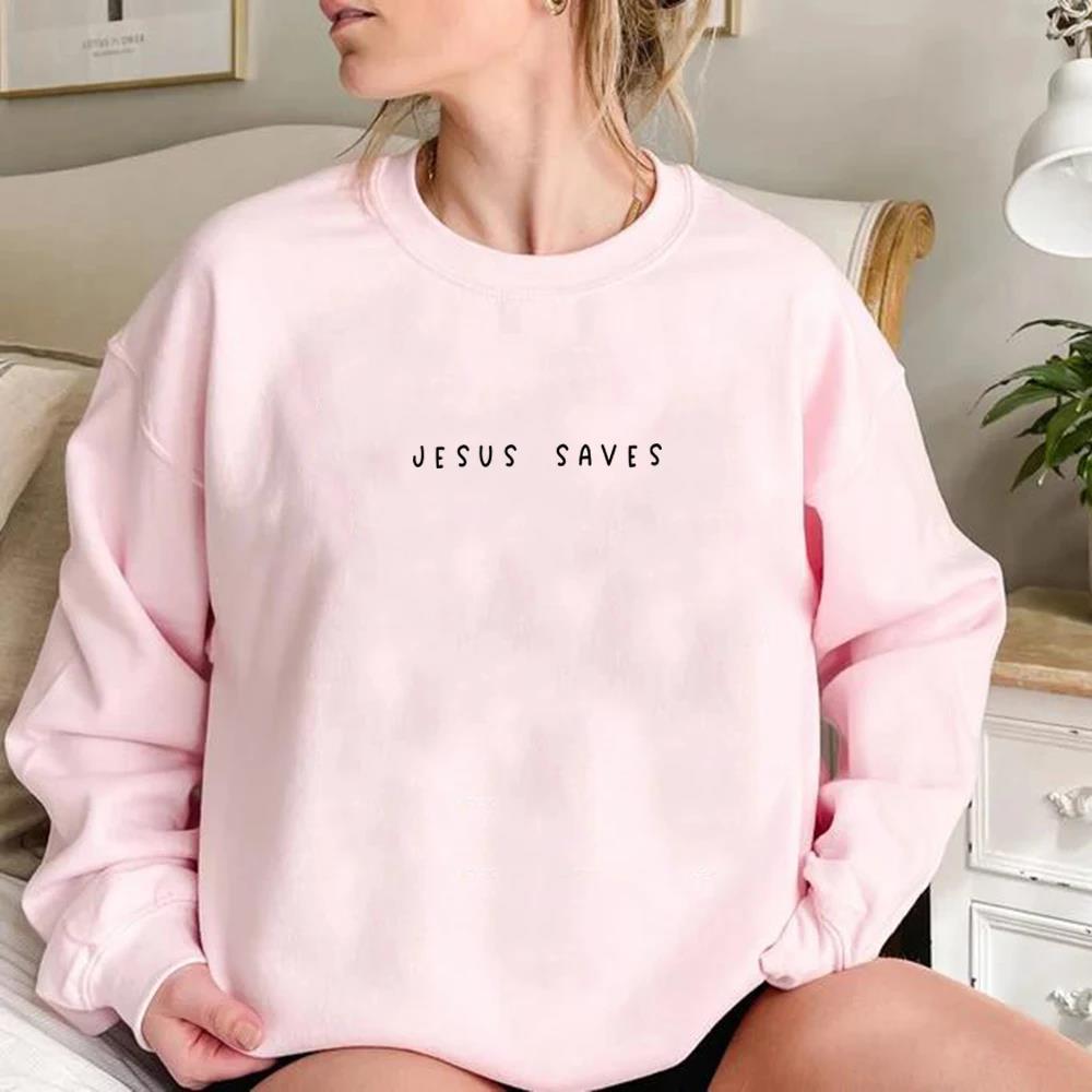 Donna seduta indossa felpa rosa con scritta "Jesus Saves", stanza accogliente, arredamento chiaro. Moda casual, abbigliamento religioso, tendenze 2023.