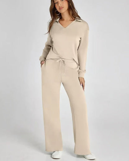 Donna in tuta beige, maglia con scollo a V e pantaloni larghi, posa su sfondo grigio. Abbigliamento casual elegante, moda donna autunno.
