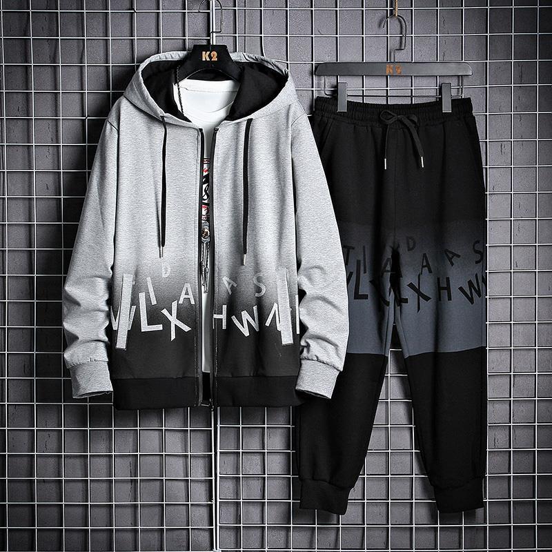 Tuta sportiva uomo grigia e nera con cappuccio, zip e pantaloni, design moderno con lettere stampate, appesa su griglia metallica.