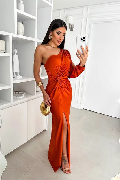 Donna in abito arancione monospalla con spacco, scatta selfie con smartphone. Elegante look da sera, accessori oro, interni moderni.