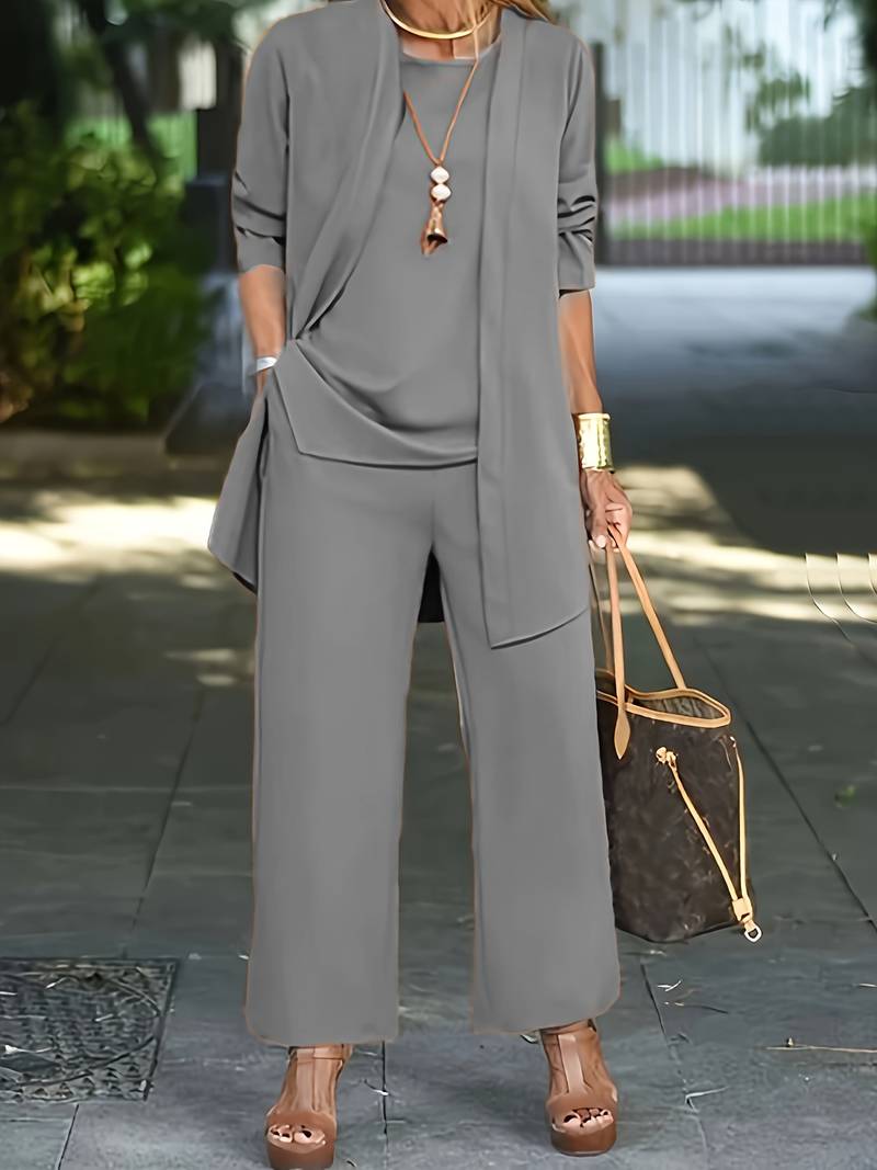 Donna in elegante completo grigio, pantaloni larghi e giacca lunga, con borsa di lusso marrone e sandali con zeppa, in contesto urbano.