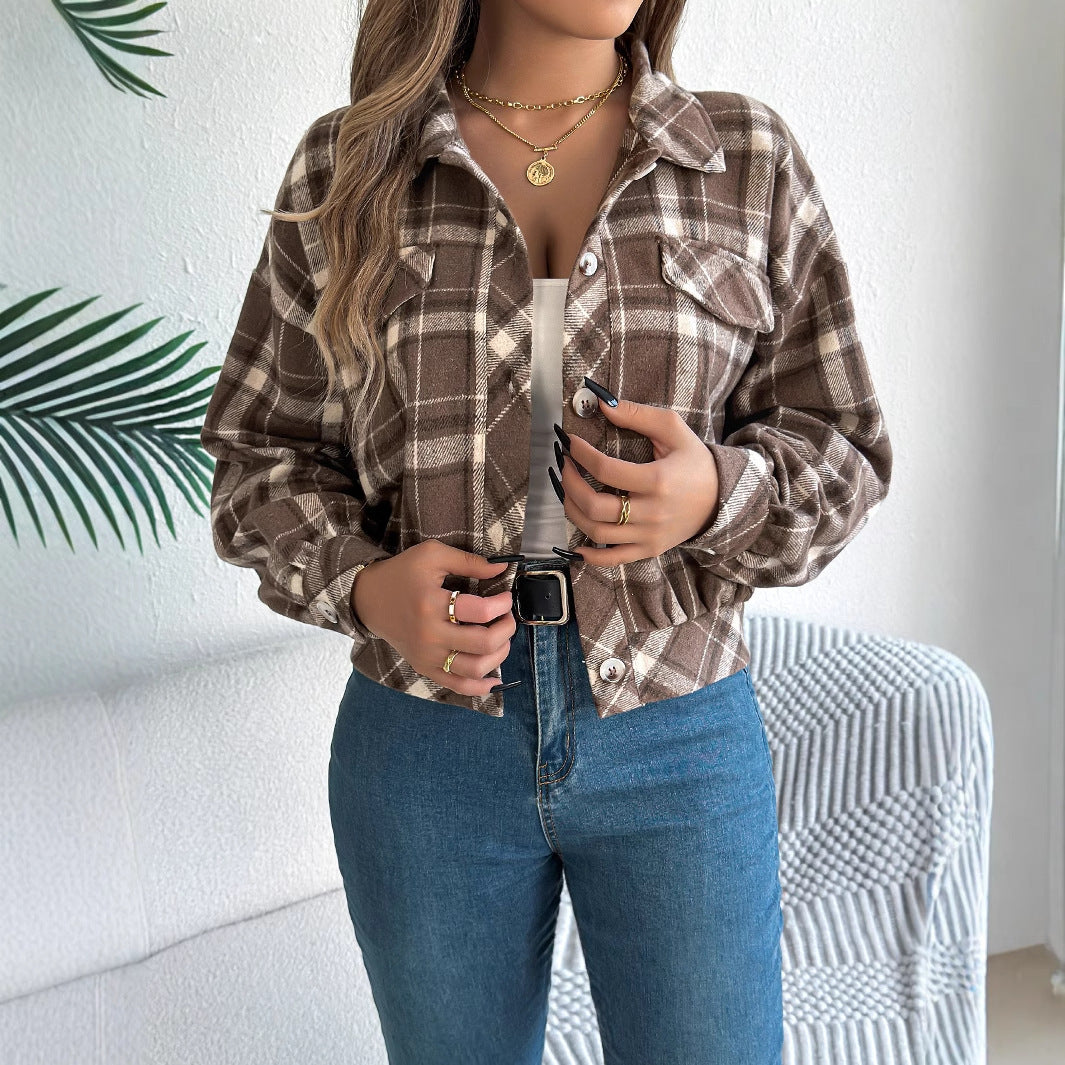 Donna con camicia a quadri marrone e jeans, indossa collana e anelli dorati. Sfondo con foglia verde e divano bianco. Moda autunnale casual.