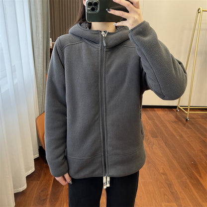 Giacca in pile grigio con cappuccio, chiusura a zip, indossata da una persona. Interno stanza con pavimento in legno e appendiabiti dorato.