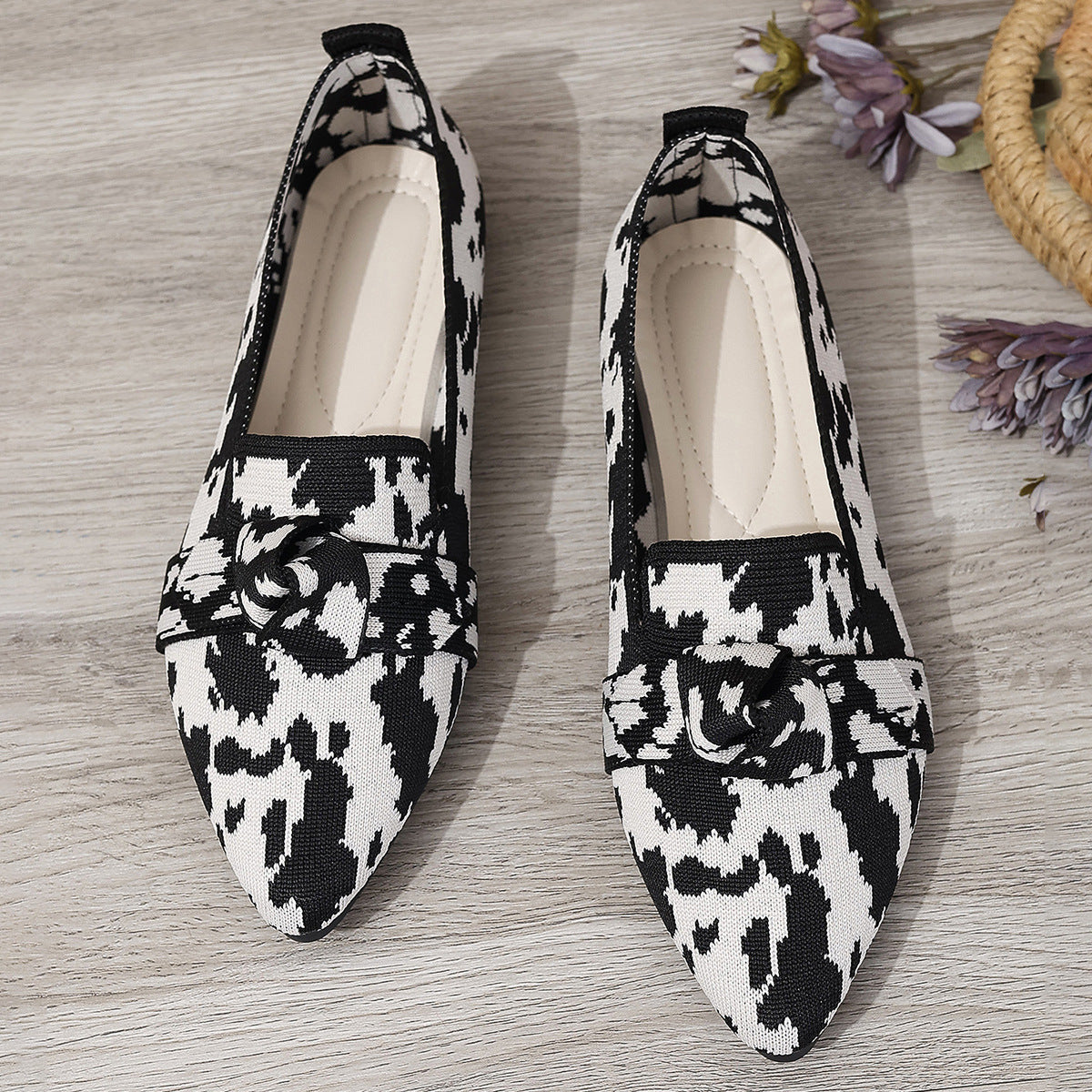 Scarpe ballerine a punta in tessuto bianco e nero con motivo astratto, su pavimento in legno chiaro, accanto a fiori secchi. Moda donna elegante.