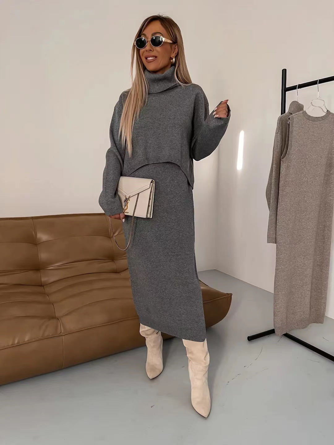 Donna con abito grigio a maglia, stivali bianchi e occhiali da sole, in un salotto moderno con divano marrone. Moda autunnale elegante.