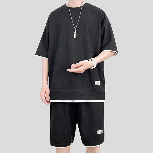 Uomo in abbigliamento casual nero, indossa t-shirt oversize e pantaloncini coordinati, con collana e orologio. Sfondo grigio chiaro.