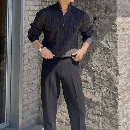 Uomo in maglione nero a coste e pantaloni neri, posa contro un muro di mattoni grigi. Moda maschile elegante, abbigliamento autunnale.