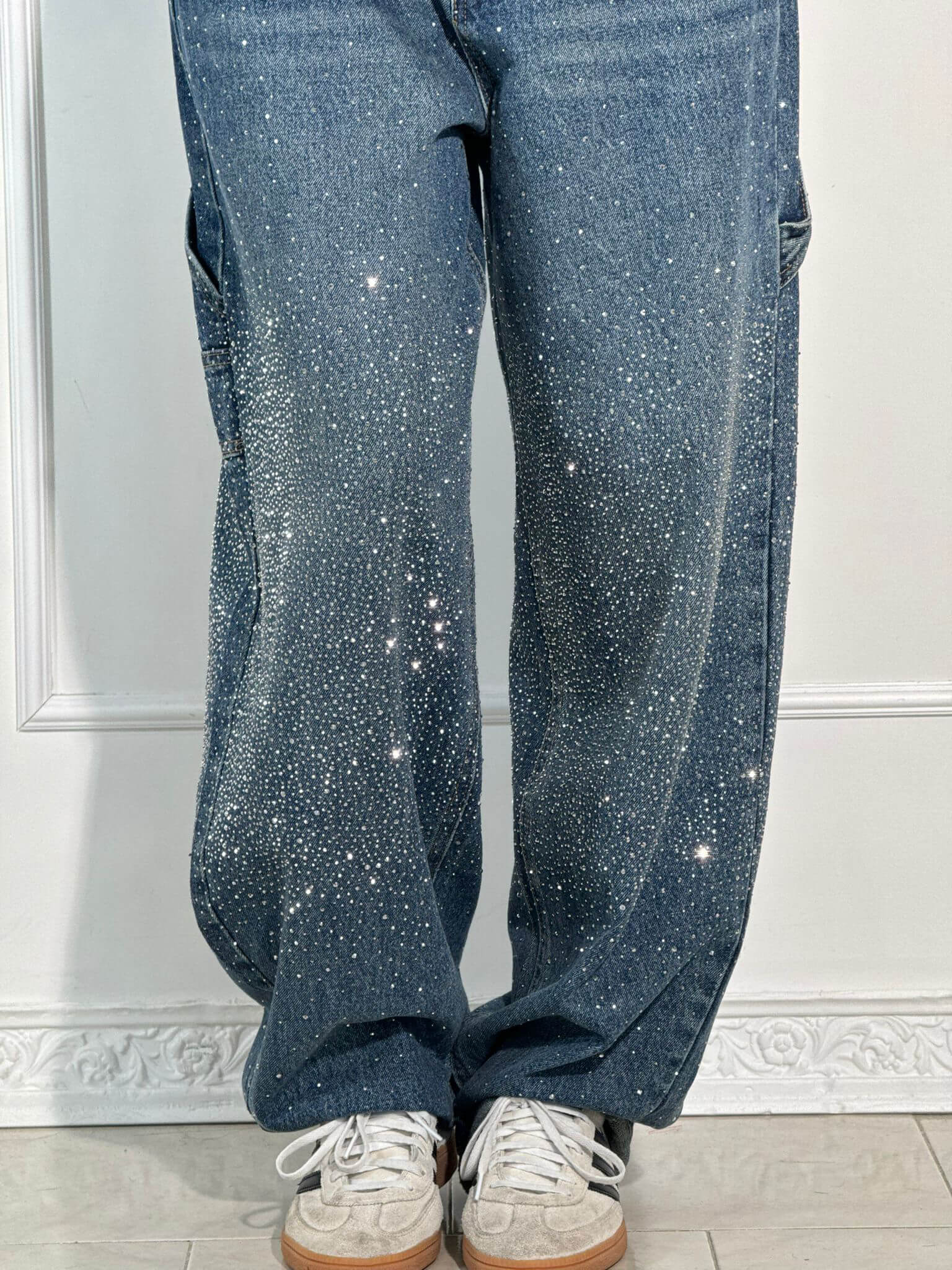 Jeans blu con strass scintillanti, abbinati a scarpe da ginnastica bianche e grigie, su sfondo bianco. Moda casual, stile glamour.