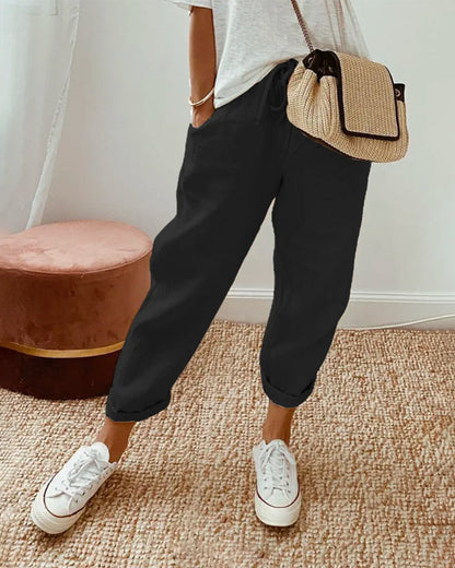 Donna con pantaloni neri casual, borsa a tracolla beige e sneakers bianche, in piedi su tappeto beige. Moda casual femminile, stile urbano.