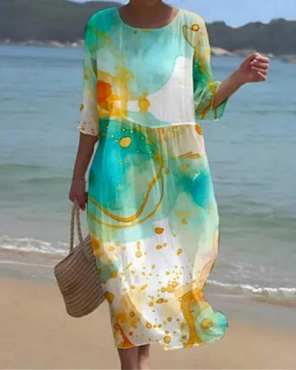 Abito lungo estivo colorato con motivi astratti verdi e arancioni, indossato da una donna su una spiaggia. Moda mare, abbigliamento estivo elegante.