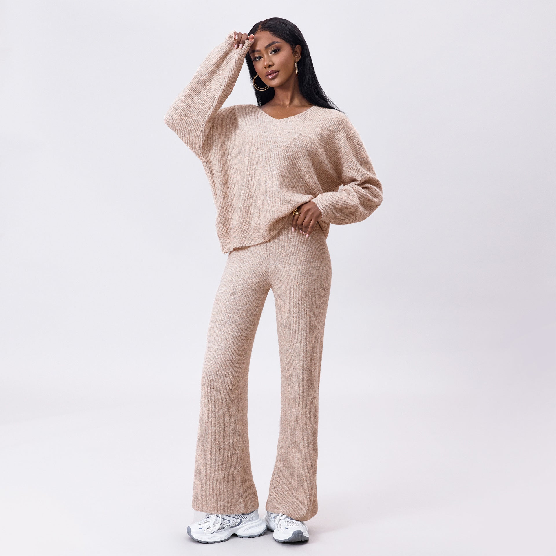 Donna in abbigliamento casual beige, maglione e pantaloni larghi, posa su sfondo bianco. Moda autunnale, stile confortevole, sneakers bianche.
