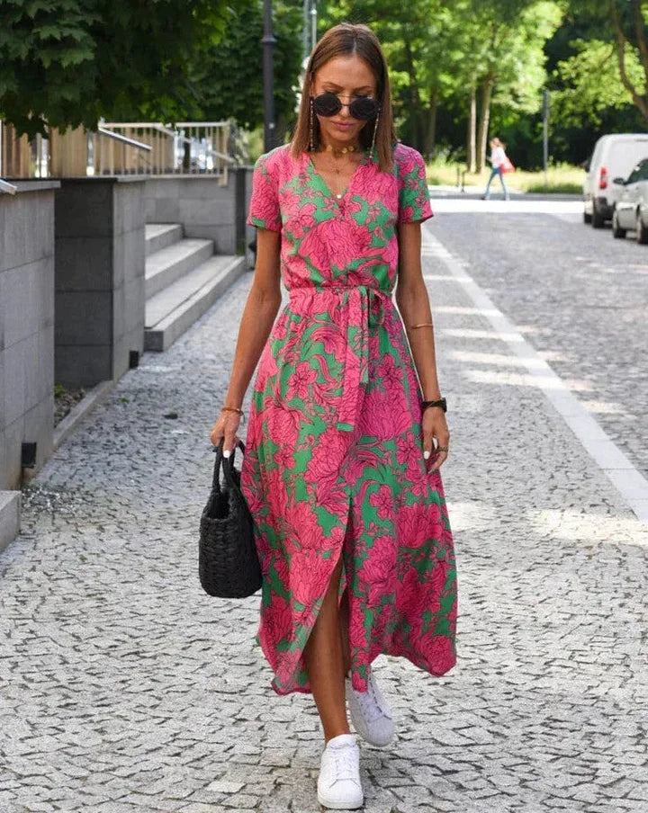 Donna con abito floreale rosa e verde, cammina su strada lastricata. Indossa occhiali da sole e scarpe bianche, borsa nera a mano.