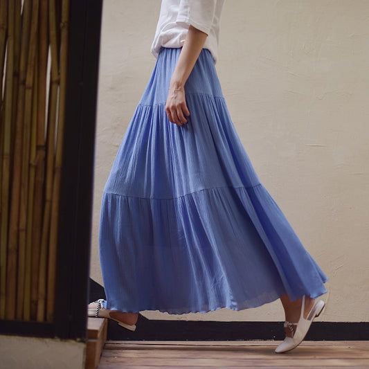 Donna con gonna lunga blu plissettata, camicia bianca e scarpe bianche, cammina su pavimento in legno. Moda estiva elegante e casual.