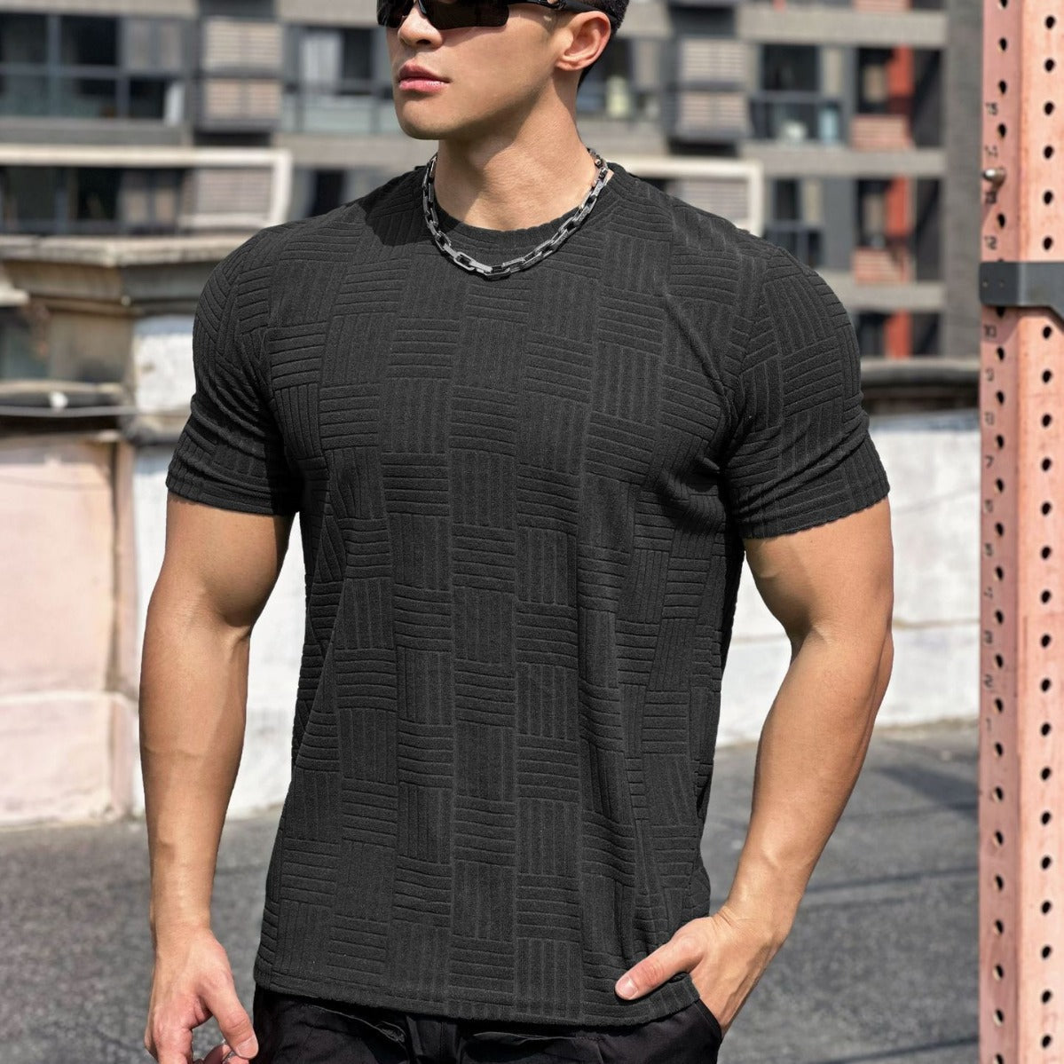 Uomo muscoloso con occhiali da sole indossa maglietta nera a trama geometrica, collana a catena, sfondo urbano. Moda maschile, stile moderno.