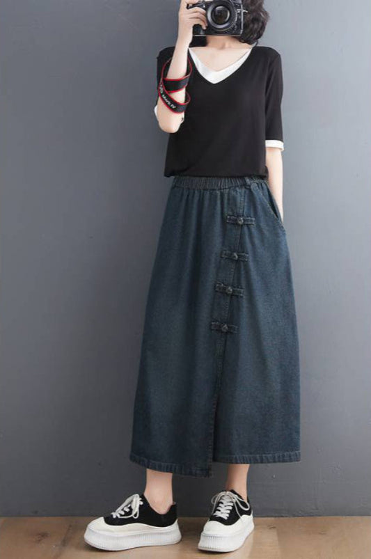 Donna con macchina fotografica indossa maglietta nera, gonna lunga in denim con bottoni decorativi e sneakers bianche e nere. Moda casual femminile.