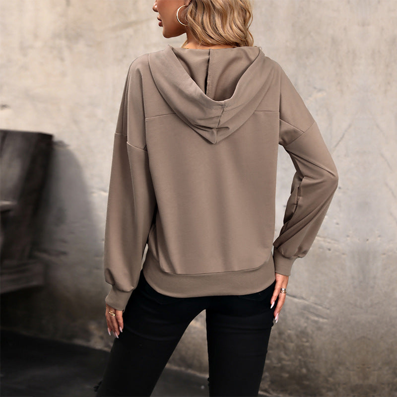 Donna di spalle con felpa beige con cappuccio, pantaloni neri, sfondo grigio. Moda casual autunnale, abbigliamento comodo e trendy.