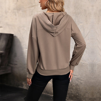 Donna di spalle con felpa beige con cappuccio, pantaloni neri, sfondo grigio. Moda casual autunnale, abbigliamento comodo e trendy.