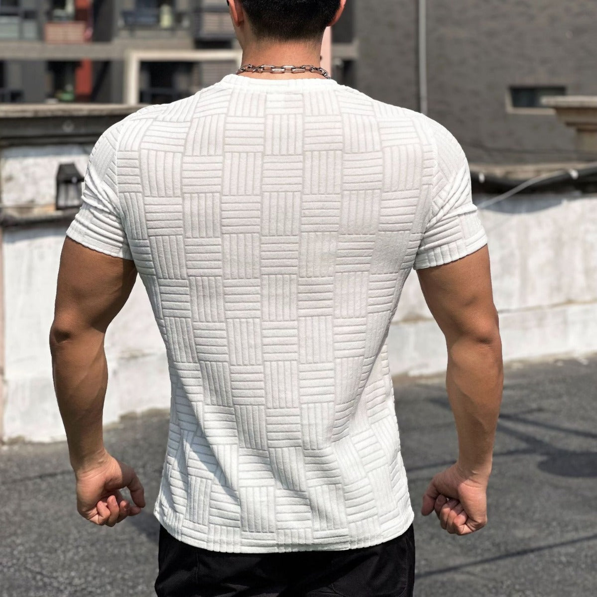 Uomo muscoloso in maglietta bianca a trama geometrica, visto di spalle su terrazza urbana. Moda maschile, abbigliamento casual estivo.