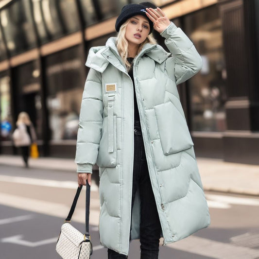 Donna con cappotto lungo imbottito verde menta, berretto nero e borsa a tracolla bianca, cammina in città. Moda invernale, street style urbano.