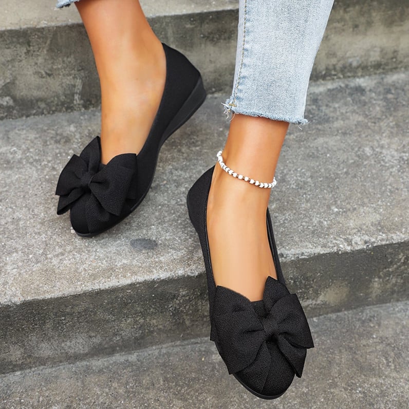 Scarpe ballerine nere con fiocco grande, indossate su gradini di cemento. Moda donna, calzature eleganti, jeans chiari, cavigliera perla.