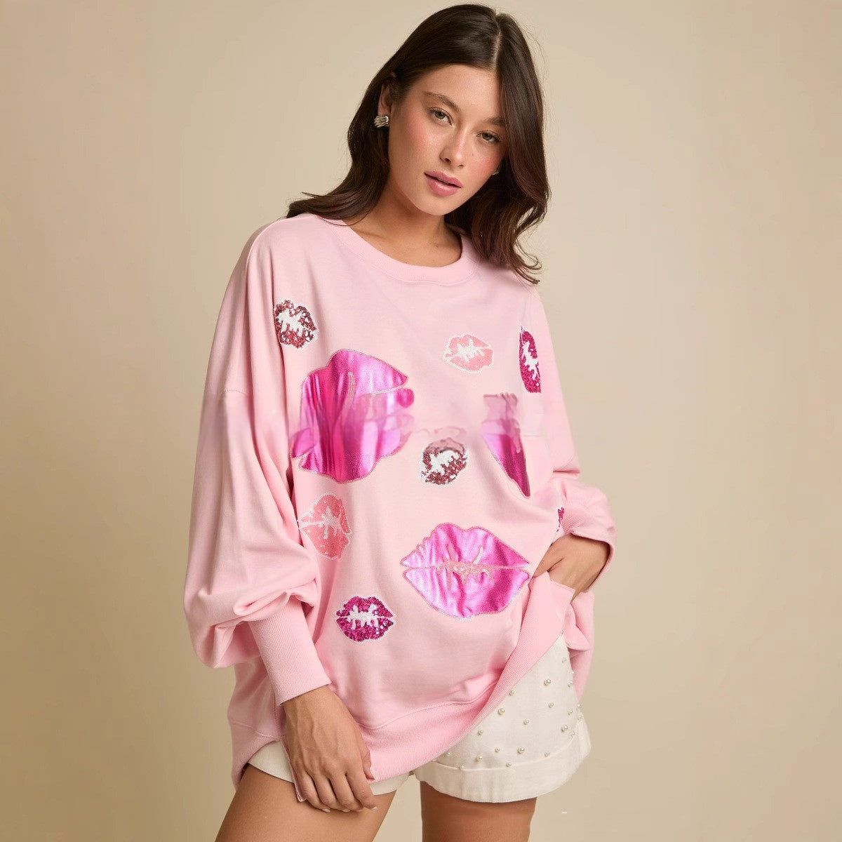 Donna con maglione rosa oversize con stampe di labbra rosa lucide, orecchini brillanti, e pantaloncini bianchi. Moda casual femminile trendy.