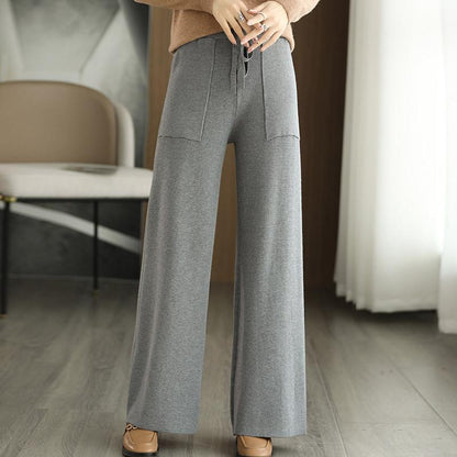 Pantaloni grigi a gamba larga, tessuto morbido, tasche frontali, indossati con scarpe marroni su pavimento in legno, ambiente moderno.