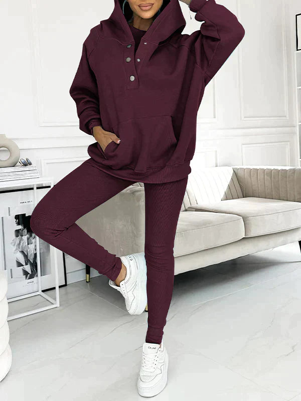 Donna in tuta sportiva bordeaux con cappuccio, posa in salotto moderno. Abbigliamento casual, moda autunnale, sneakers bianche.