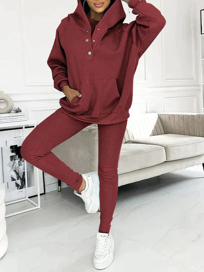 Donna in tuta sportiva rossa con cappuccio, pantaloni aderenti e sneakers bianche, posa in salotto moderno. Abbigliamento casual, moda autunno.