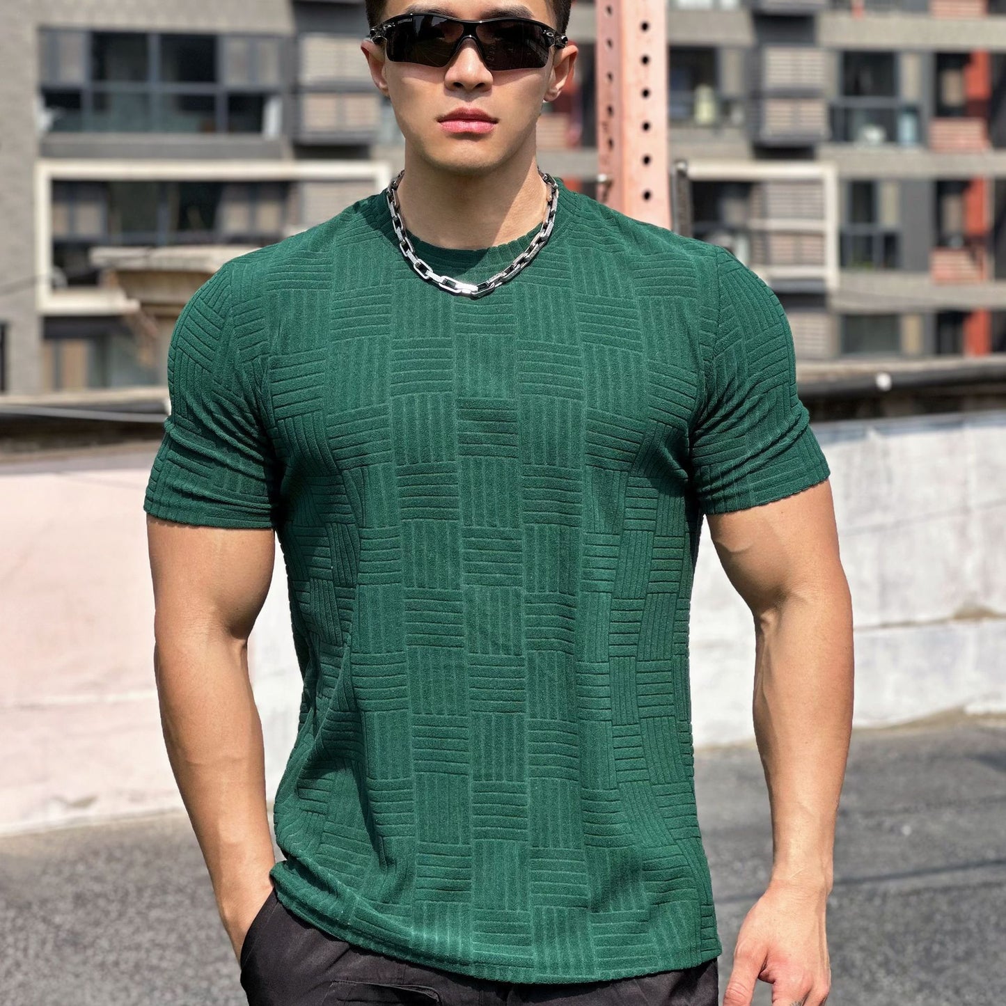 Uomo muscoloso con occhiali da sole, indossa maglietta verde a trama geometrica e catena d'argento, sfondo urbano.