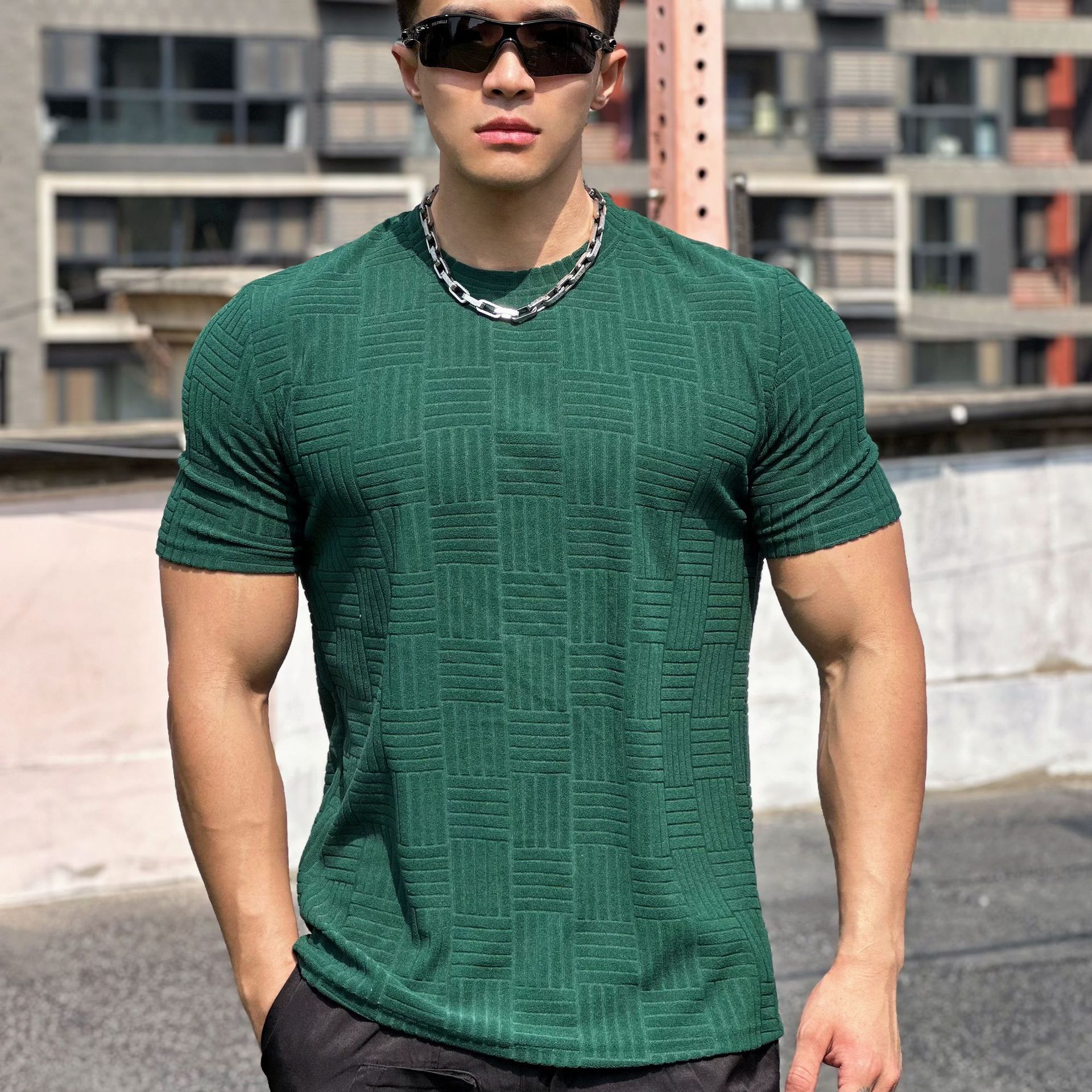 Uomo muscoloso con occhiali da sole, indossa maglietta verde a trama geometrica e catena d'argento, sfondo urbano.
