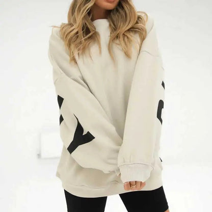 Donna con maglione oversize beige, maniche lunghe con scritte nere, abbinato a leggings neri. Moda casual autunnale, stile confortevole.