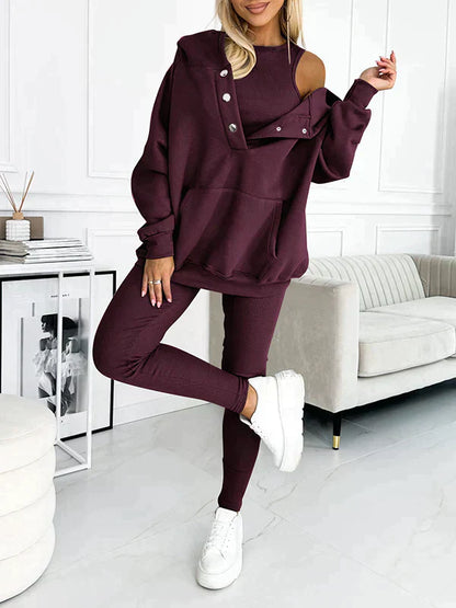Donna in tuta sportiva bordeaux con spalla scoperta, felpa con bottoni e pantaloni abbinati, posa in salotto moderno con divano chiaro e decorazioni minimal.