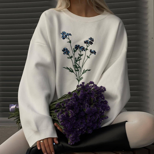 Donna con maglione bianco decorato con fiori blu, tiene un mazzo di fiori viola. Moda autunnale, stile casual chic, tendenze abbigliamento 2023.
