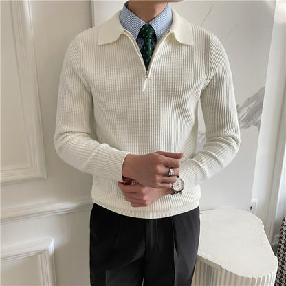 Uomo in maglione bianco a collo alto con zip, camicia azzurra e cravatta verde, indossa orologio e anelli, posa elegante in ambiente interno.