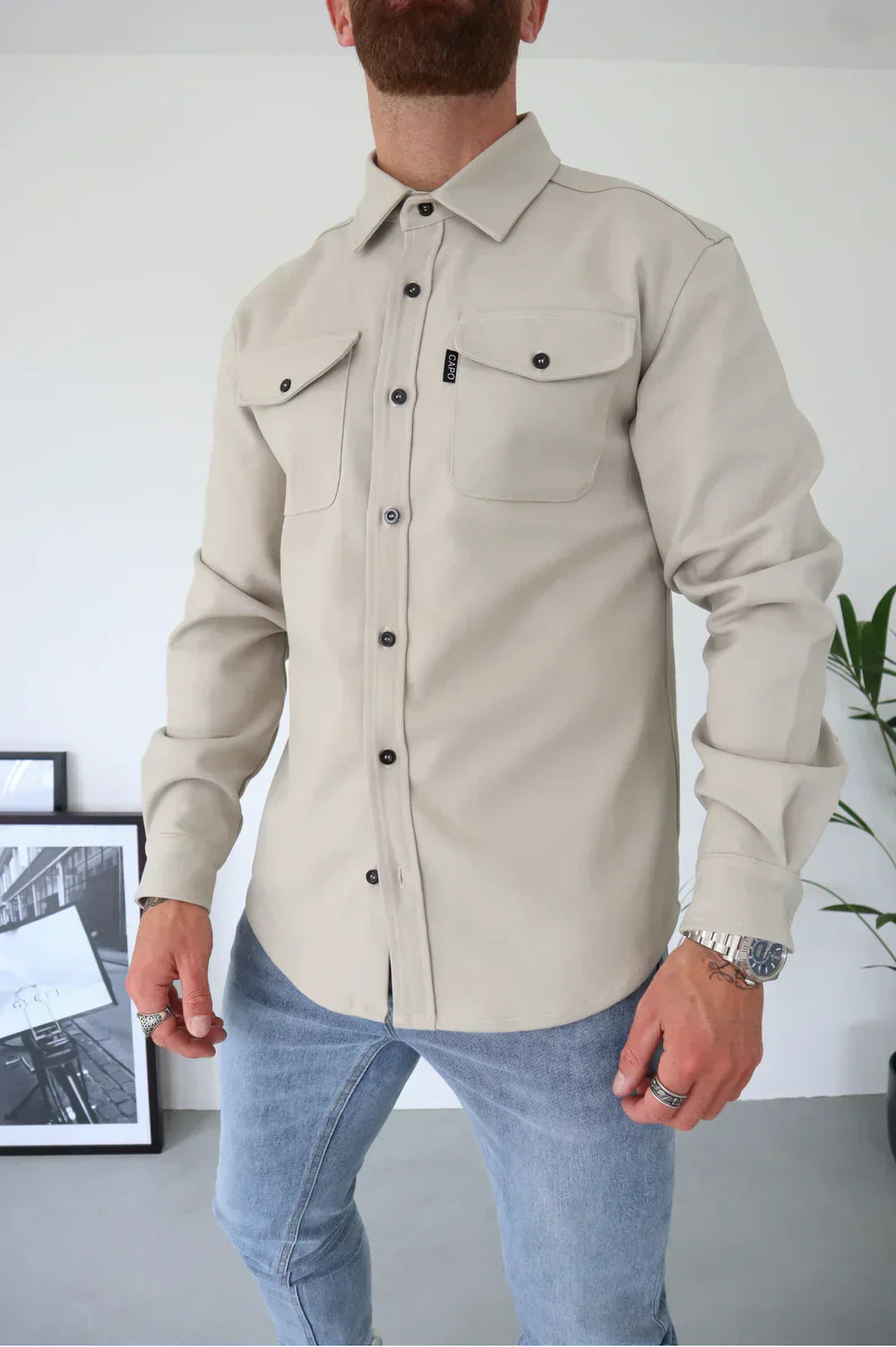 Camicia beige da uomo con tasche frontali e bottoni neri, abbinata a jeans chiari. Look casual ed elegante, ideale per moda maschile contemporanea.