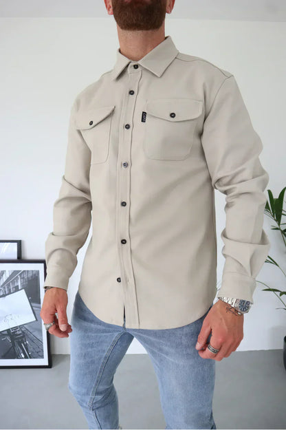 Camicia beige da uomo con tasche frontali e bottoni neri, abbinata a jeans chiari. Look casual ed elegante, ideale per moda maschile contemporanea.