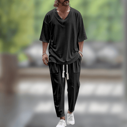 Uomo con abbigliamento casual nero, maglietta e pantaloni larghi, cammina in un ambiente moderno. Scarpe bianche, sfondo sfocato verde.