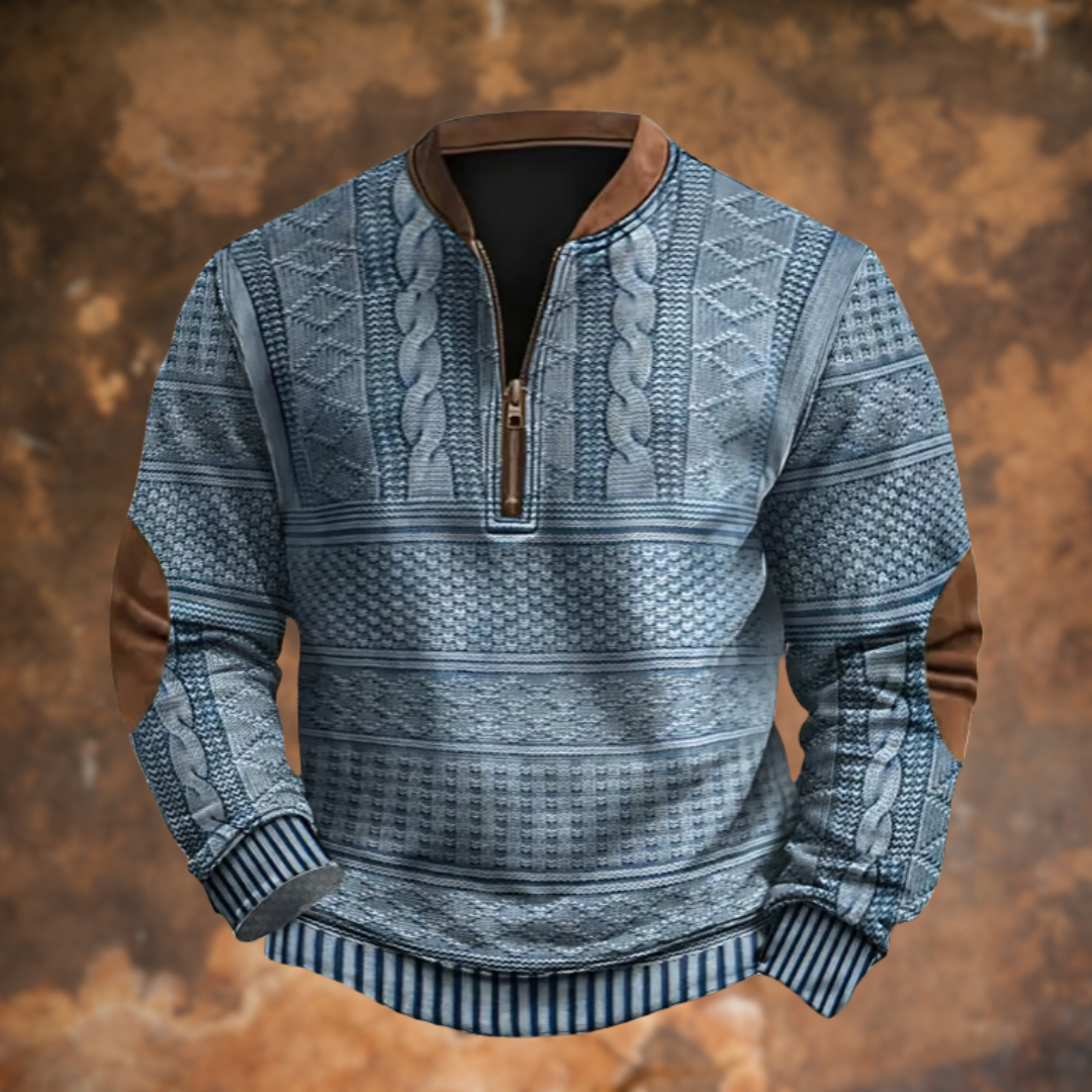 Maglione invernale blu con motivo intrecciato, zip frontale, dettagli in pelle marrone su gomiti e colletto, sfondo marrone sfocato.