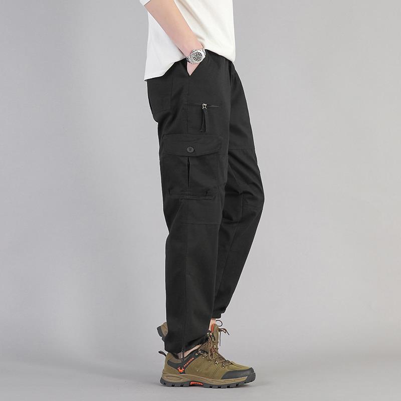 Pantaloni cargo neri da uomo, tasche multiple, stile casual, abbinati a scarpe da trekking verdi, sfondo grigio.