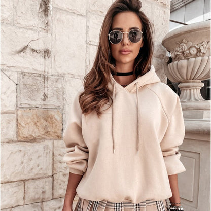 Donna con occhiali da sole indossa felpa beige e gonna a quadri, posa davanti a muro di pietra. Moda autunnale, stile casual chic.