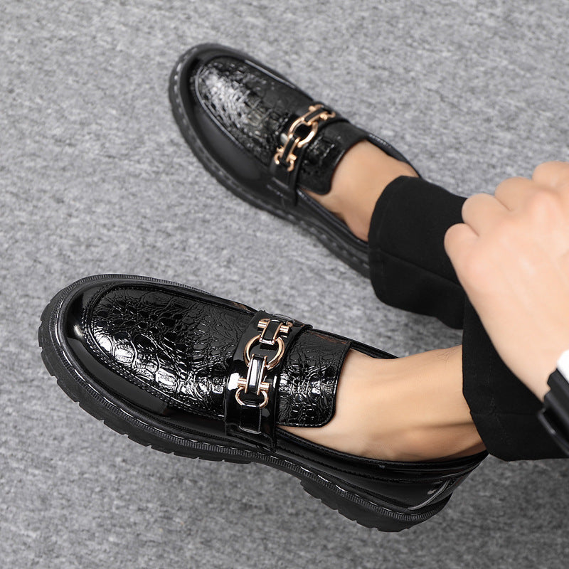 Scarpe mocassini neri lucidi con catena dorata, indossati su pavimento grigio. Eleganti e alla moda, ideali per look formali e casual.