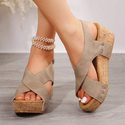 Sandali donna beige con zeppa in sughero, design incrociato, dettagli metallici, indossati su pavimento in legno. Accessorio cavigliera perle.
