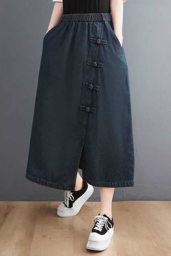 Gonna lunga in denim blu scuro con bottoni decorativi, tasche laterali, abbinata a scarpe da ginnastica bianche e nere su sfondo grigio.