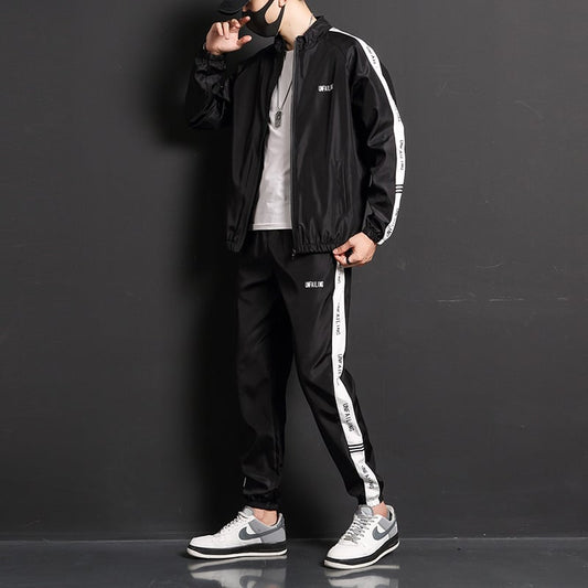 Uomo in tuta sportiva nera con strisce bianche, indossa scarpe da ginnastica grigie e cappellino nero, posa su sfondo grigio scuro.