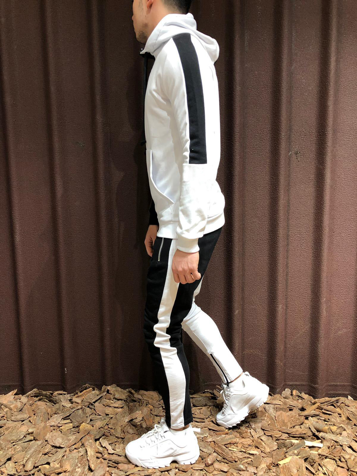 Uomo in tuta sportiva bianca e nera con cappuccio, scarpe da ginnastica bianche, su sfondo marrone. Moda casual, abbigliamento sportivo uomo.