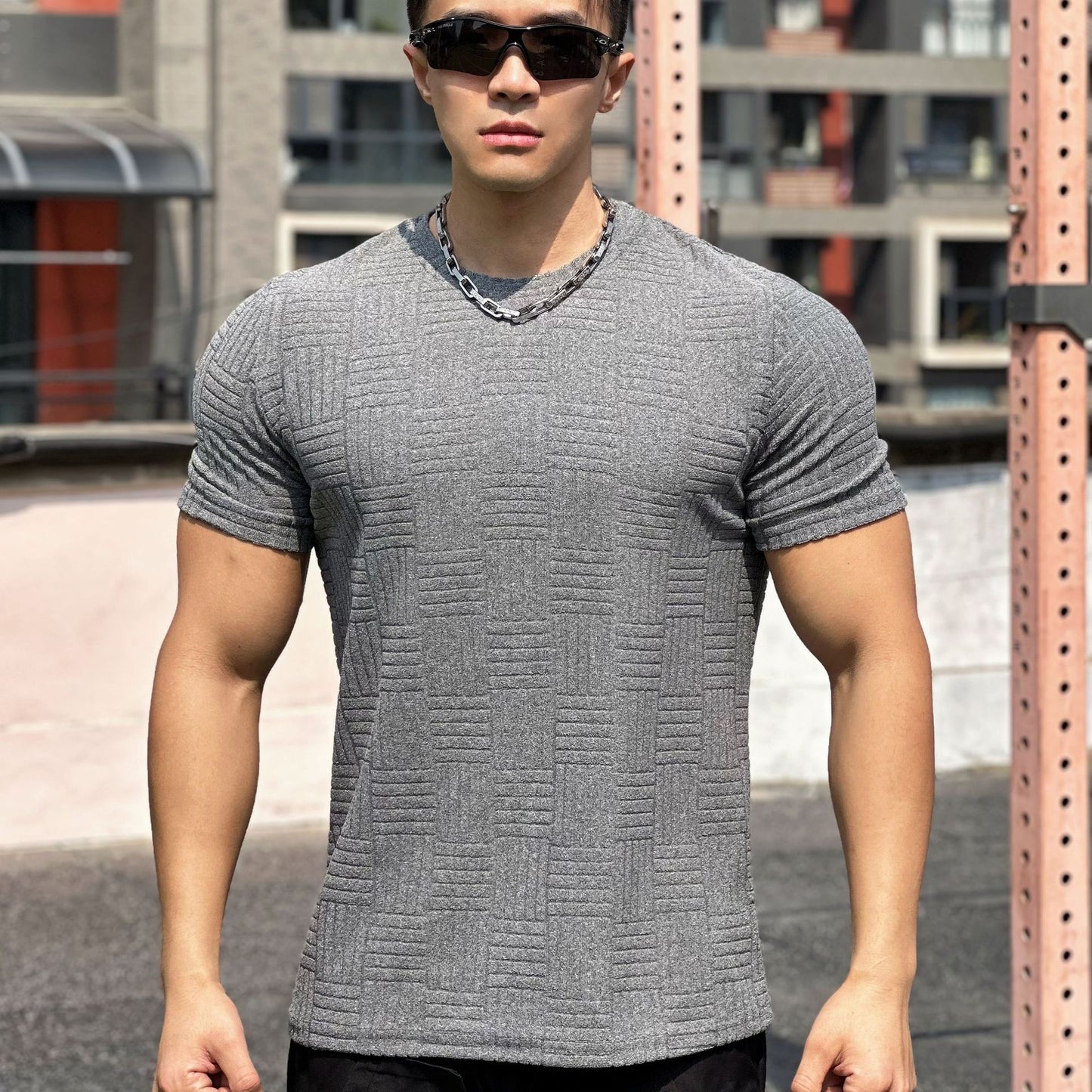 Uomo muscoloso con occhiali da sole indossa maglietta grigia a trama geometrica, posa all'aperto su sfondo urbano. Moda maschile casual.