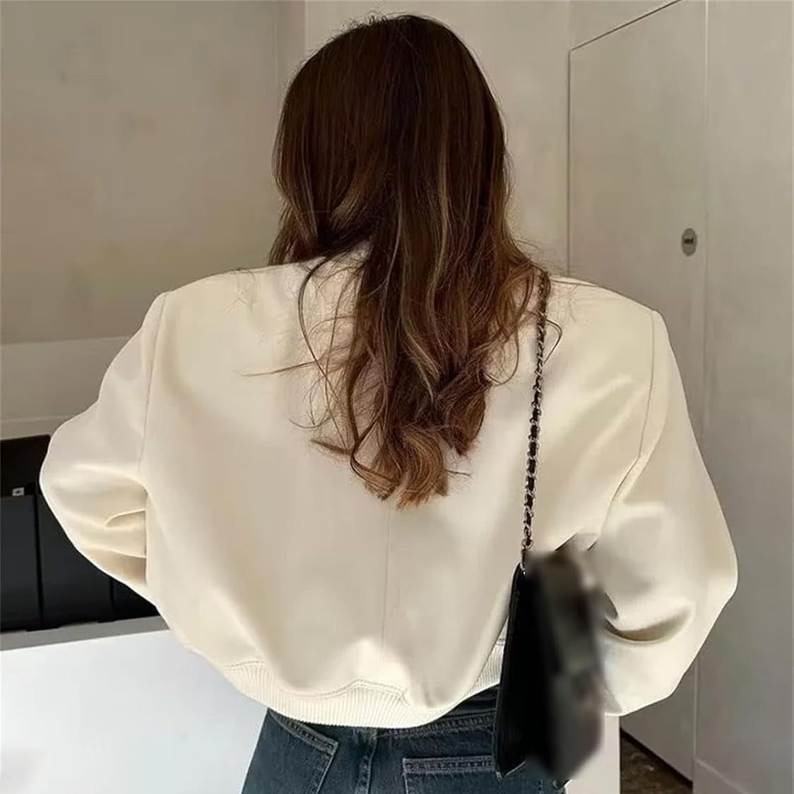 Donna di spalle con giacca bianca e borsa nera, capelli castani ondulati, in un ambiente interno moderno. Moda autunnale casual.