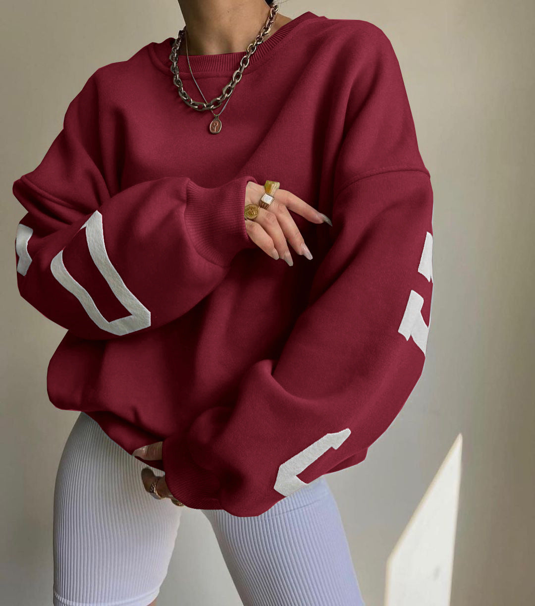 Donna con felpa oversize bordeaux con numeri bianchi, collana a catena e anelli dorati, indossa leggings bianchi. Moda casual autunno.