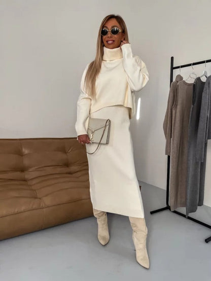 Donna con abito invernale bianco, stivali alti e occhiali da sole, accanto a divano marrone e appendiabiti con vestiti neutri. Moda elegante autunnale.